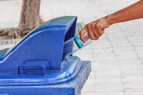 A close-up of a person's hand placing a plastic bottle into a blue public rubbish bin, situated outdoors on a paved area with light-colored tiles. The bin features a curved lid with an opening at the top and is made of durable plastic with a slightly textured surface. In the background, a small tree trunk and a portion of paved ground are visible, indicating an urban or park setting. The scene is well-lit, suggesting daytime, and the action demonstrates typical waste disposal within a city environment, relevant to commercial waste removal services in Bromley. The image emphasizes responsible rubbish disposal practices for household or commercial waste management, aligning with services offered by Commercial Waste Removal Bromley.